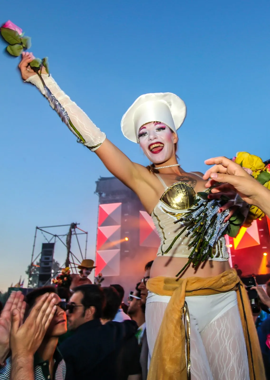 A performer in white and gold on stilts waving flowers above a festival crowd under a blue sky