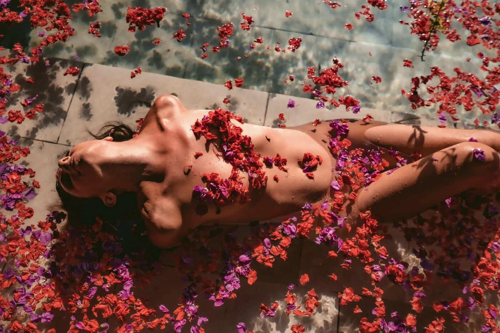 A woman lying bare in a shallow pool strewn with red and purple petals, seen from above