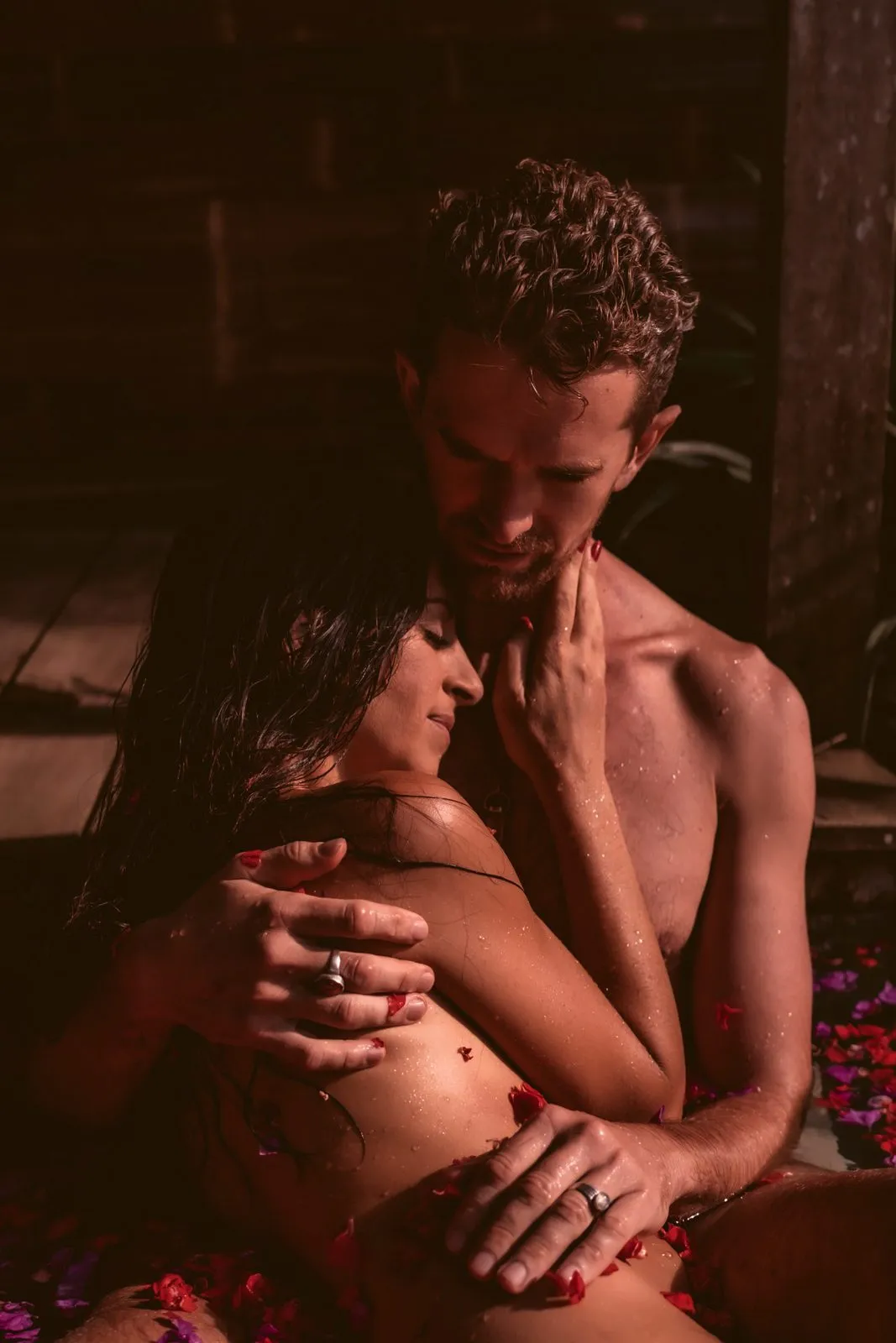 A couple holding each other close in warm light, petals on their skin