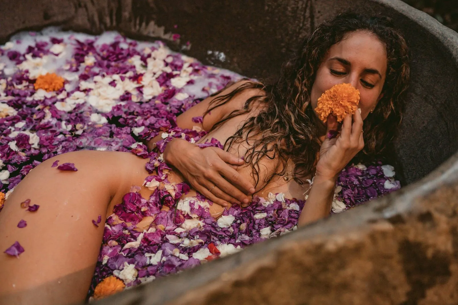 A woman in a stone bath full of petals, holding a marigold to her face with her eyes closed