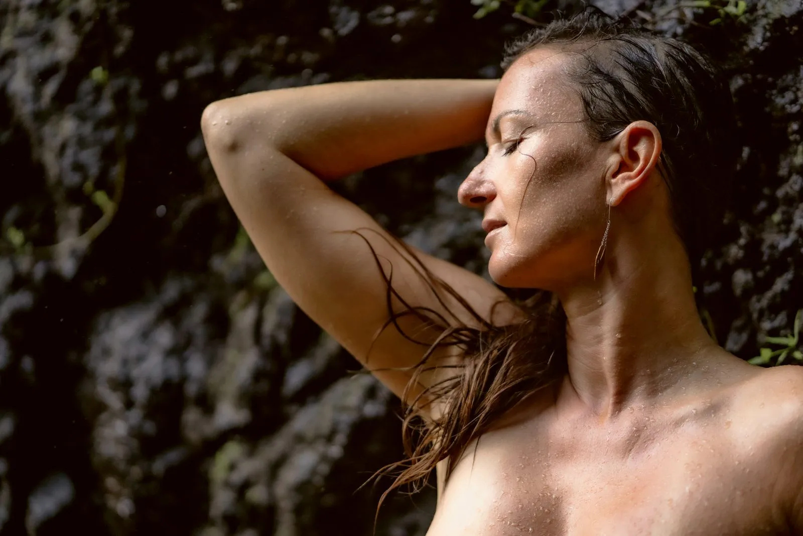 A woman with wet hair and a hand behind her head against dark rock, eyes closed in the sun