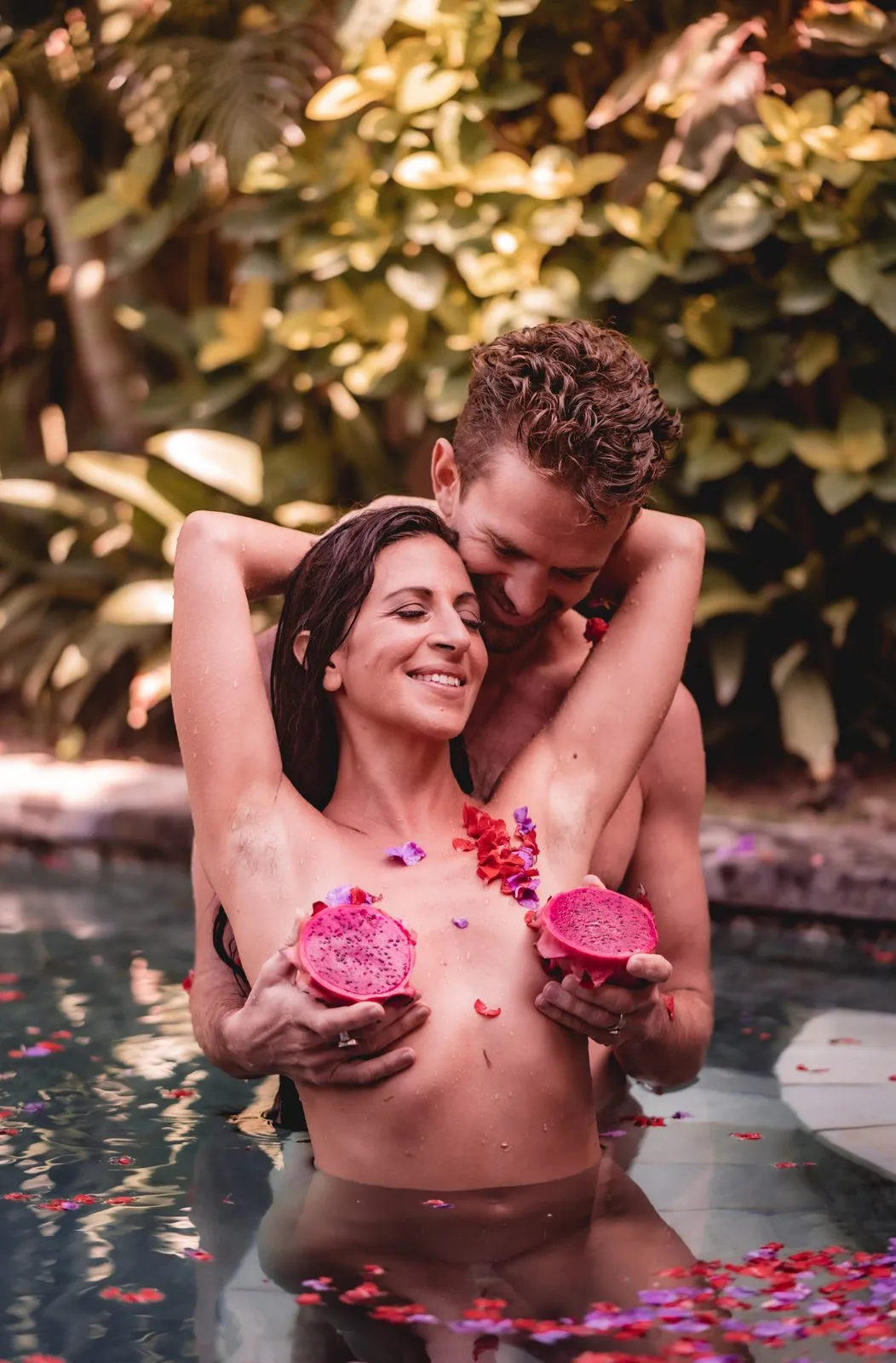 A couple in a petal-strewn pool, his arms around her from behind, each holding half a dragon fruit