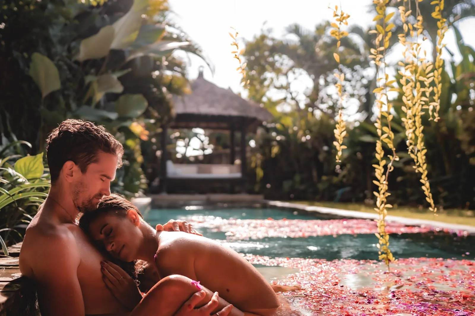 A couple resting at the edge of a villa pool covered in petals, a Balinese pavilion behind them