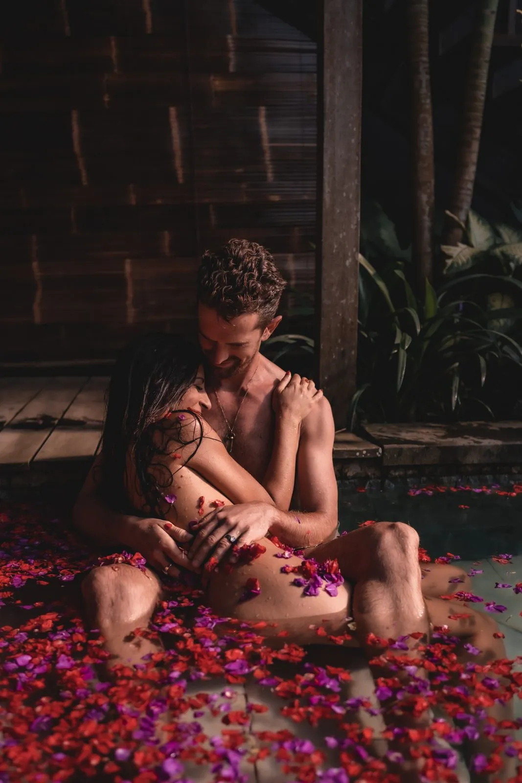 A couple in a stone bath full of red petals, holding each other, a wooden hut behind