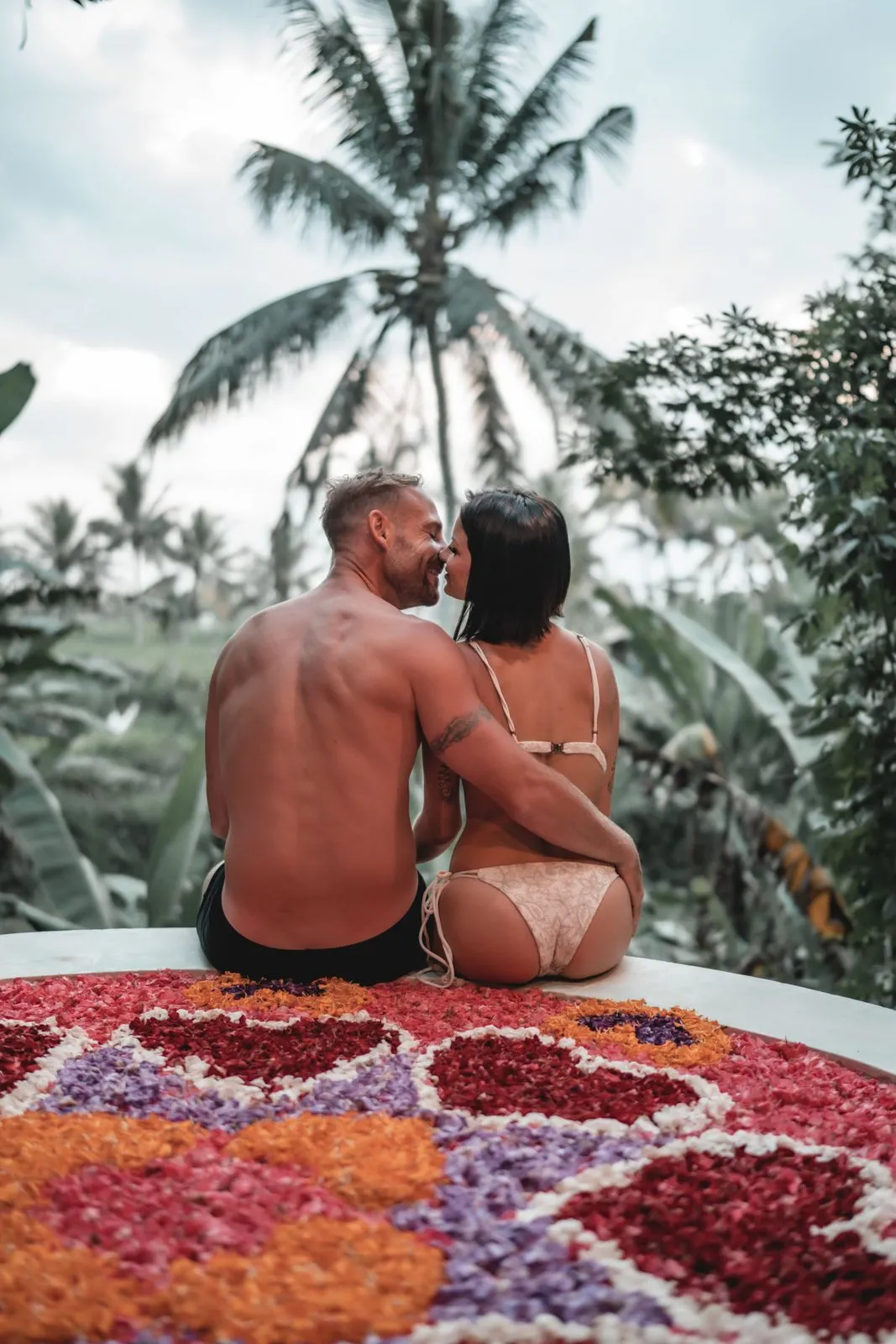 A couple sitting close on a round bed of petals arranged in a heart, looking out over palm trees