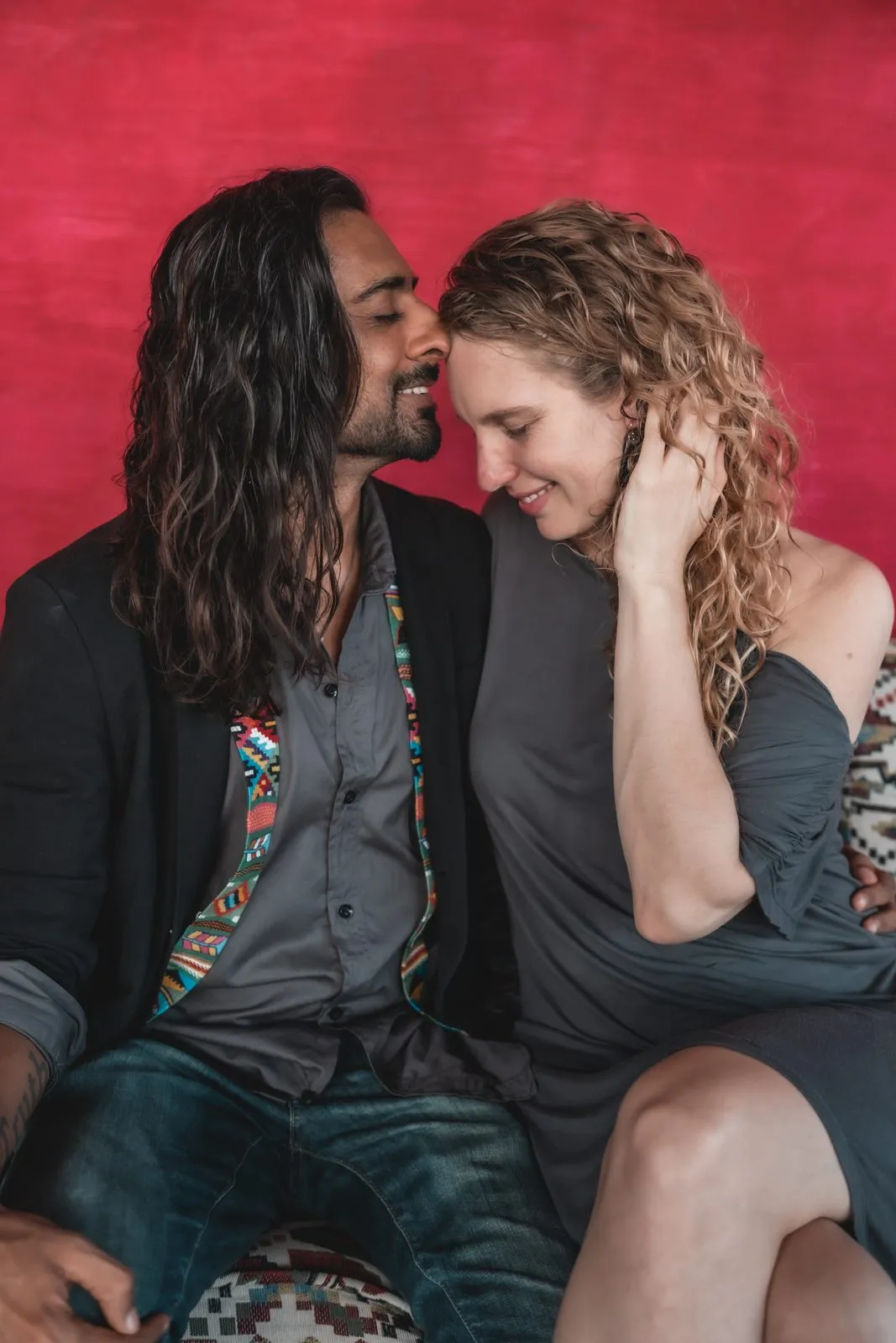 A couple sitting close against a red wall, nose to nose and laughing