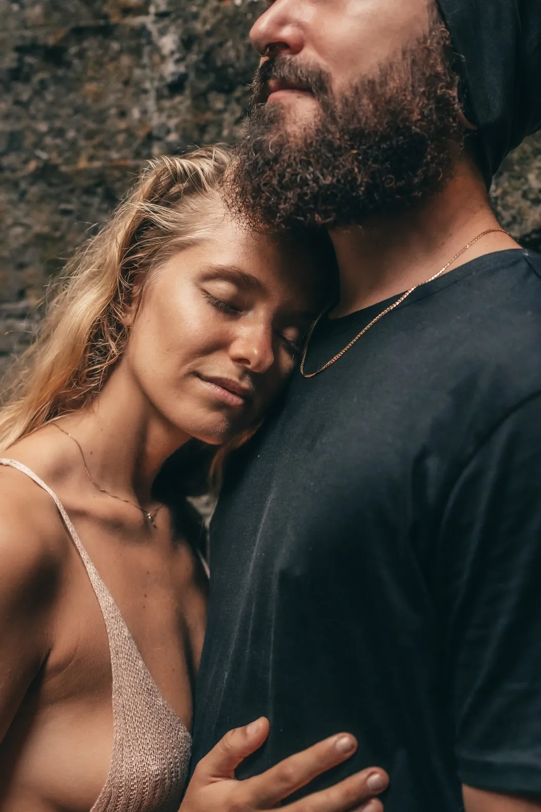 A woman resting her face against a bearded man's chest, eyes closed, before an old stone wall