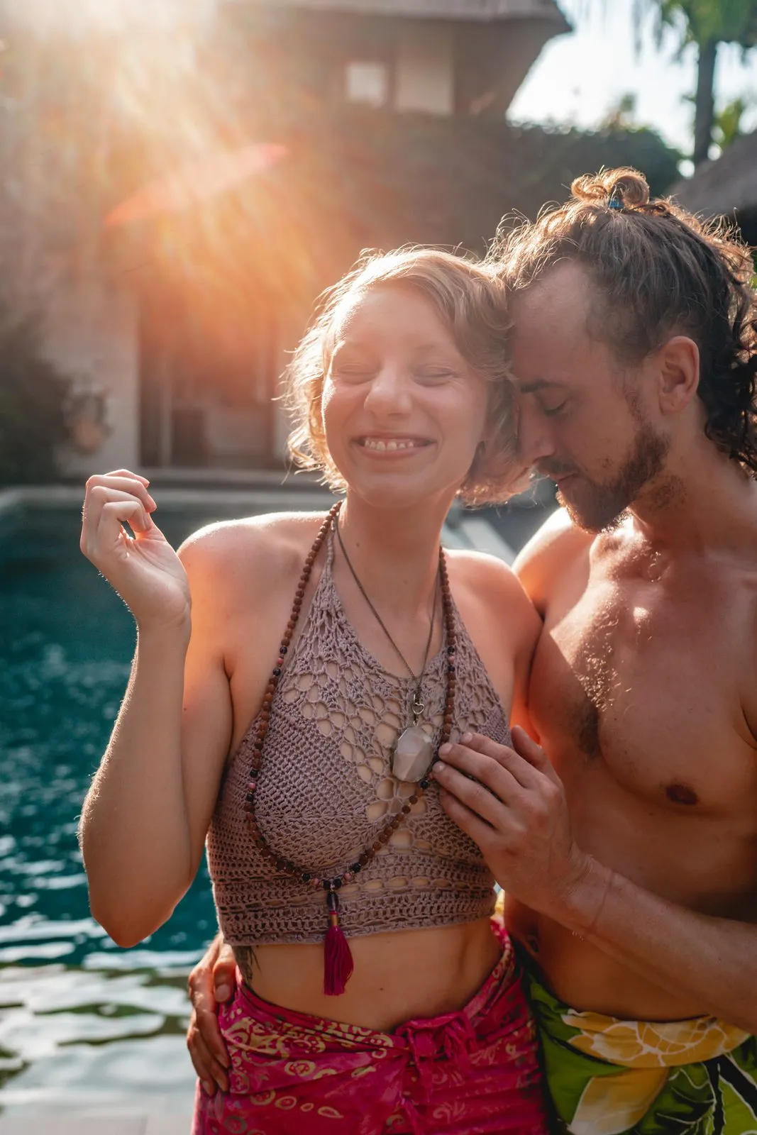 A couple laughing by a pool in the sun, she in a beaded top and red sarong