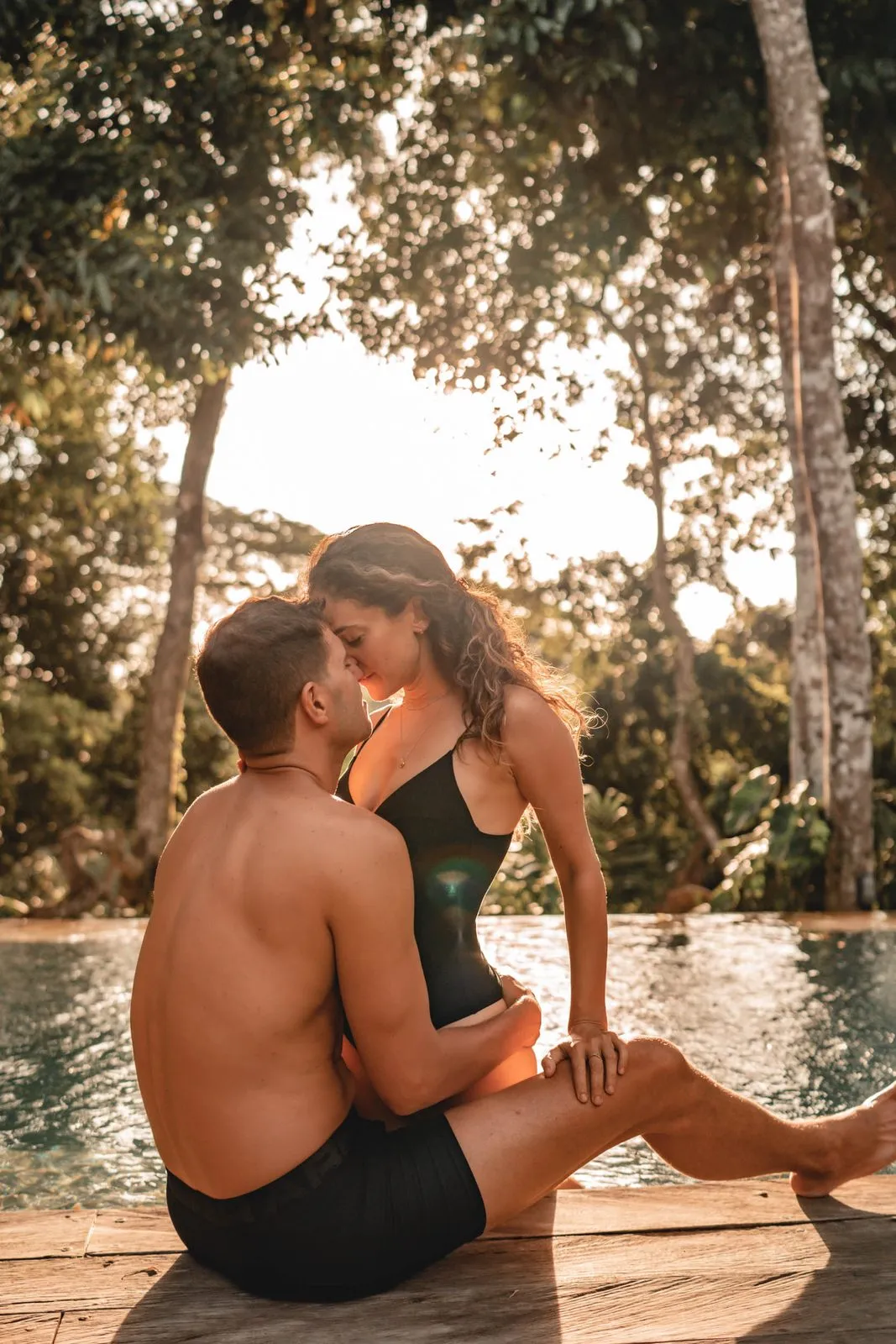 A couple sitting at the edge of an infinity pool at golden hour, her on his lap, forehead to forehead