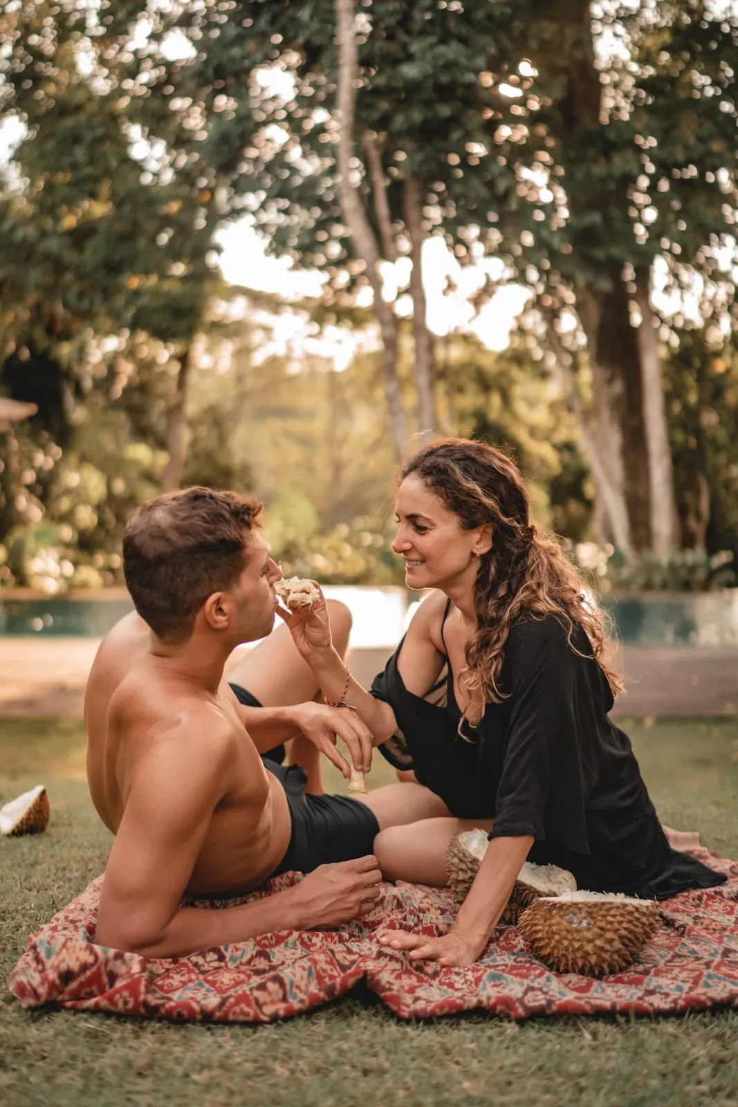 A couple laughing on a picnic rug on the grass, a pool and tall trees behind them