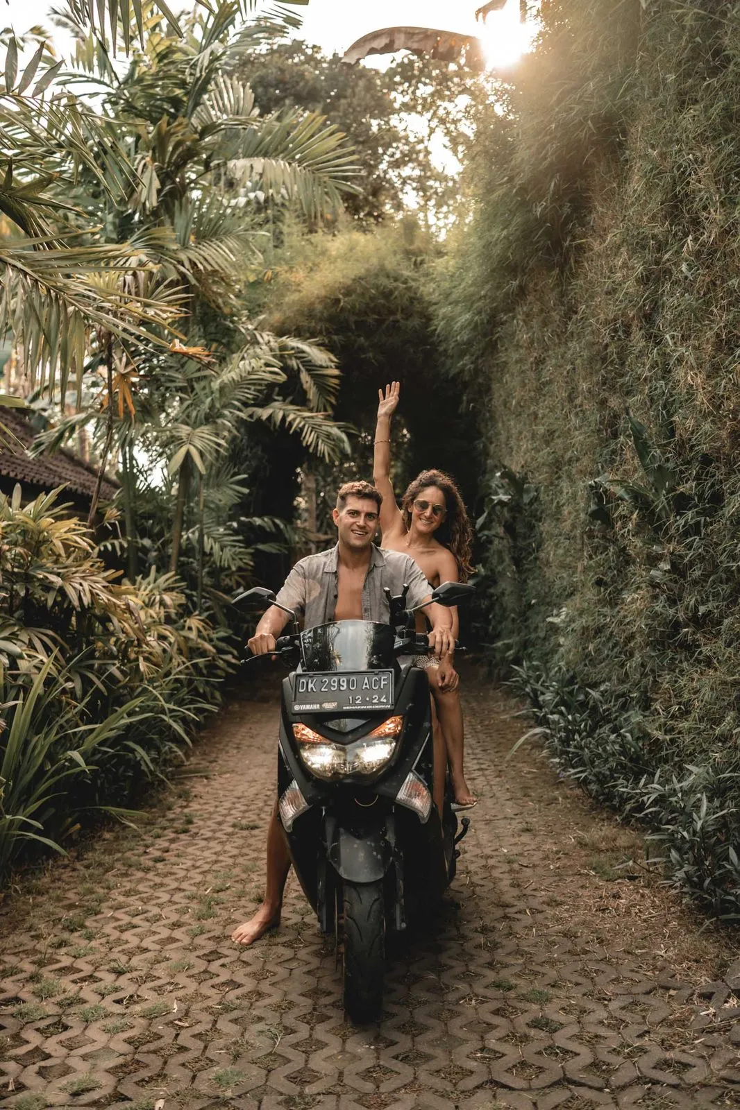 A couple on a scooter in a narrow green lane in Bali, her arm raised in the air