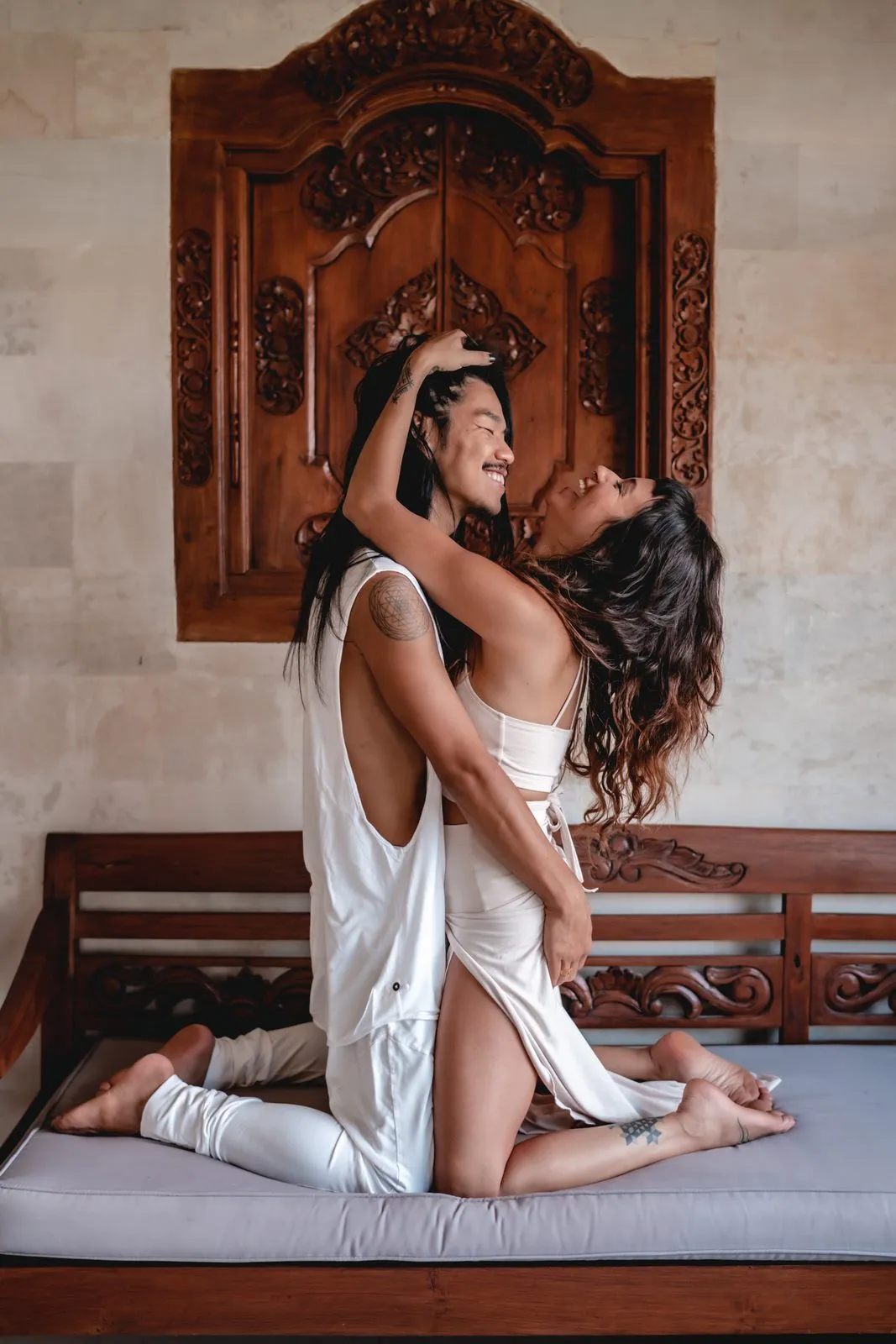 Two women in white embracing on a daybed before a carved Balinese door