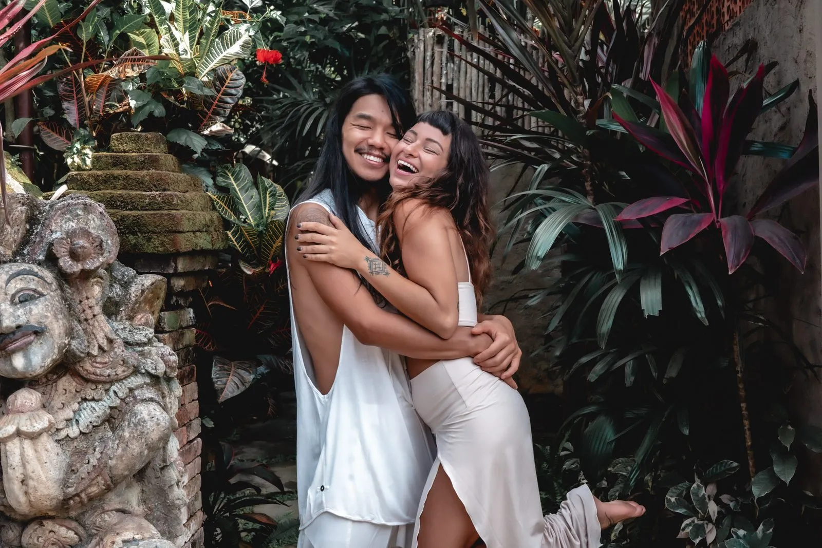 Two women in white laughing in a hug in a tropical garden, a stone statue beside them
