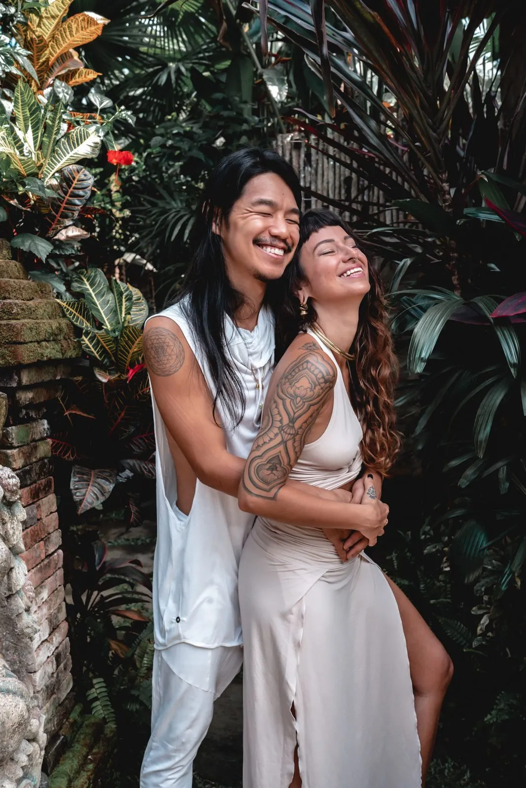 A couple in white laughing together in a leafy Balinese garden, his arm around her shoulder