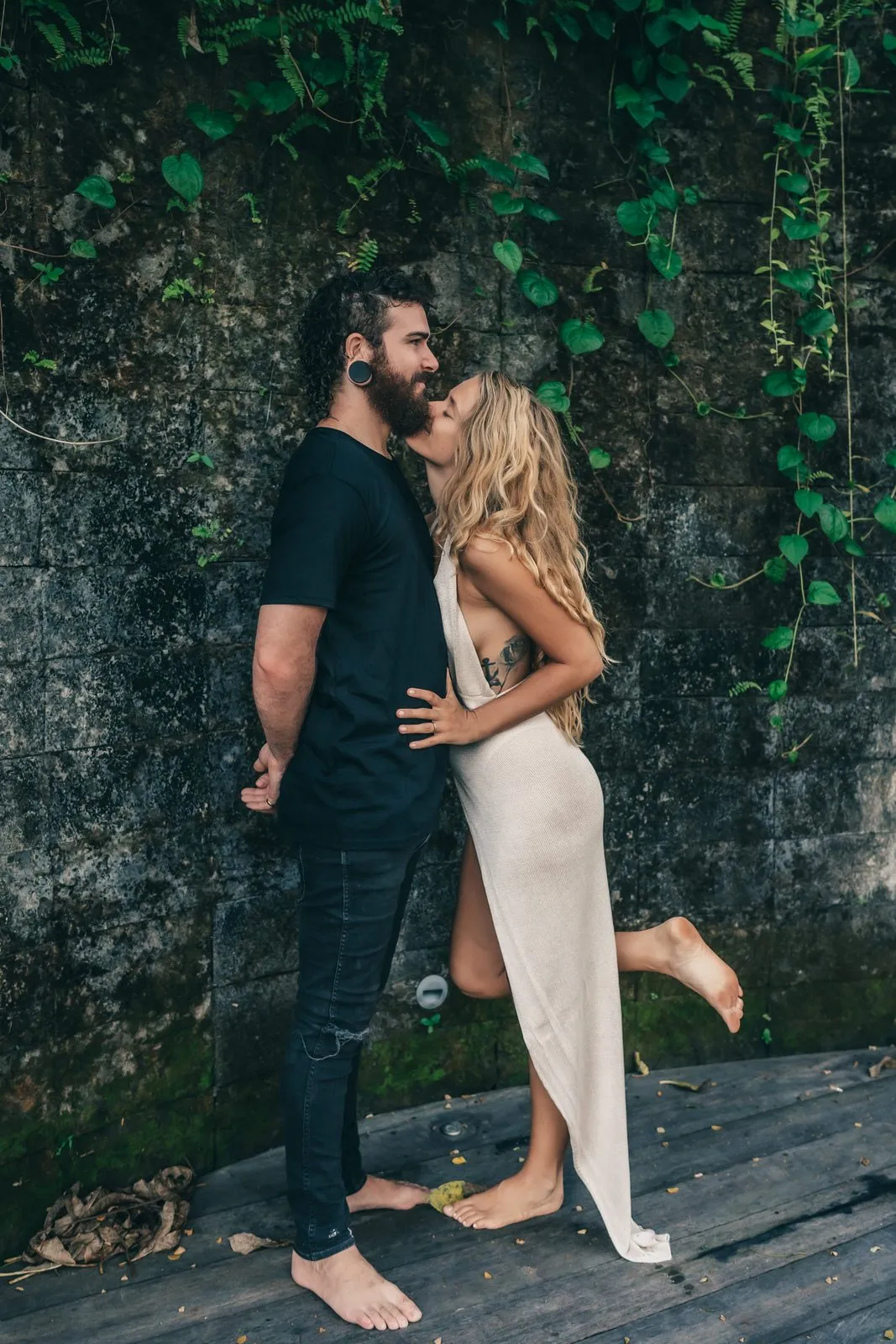 A couple embracing on a wooden deck before an ivy-covered wall, she in a cream dress, one foot lifted