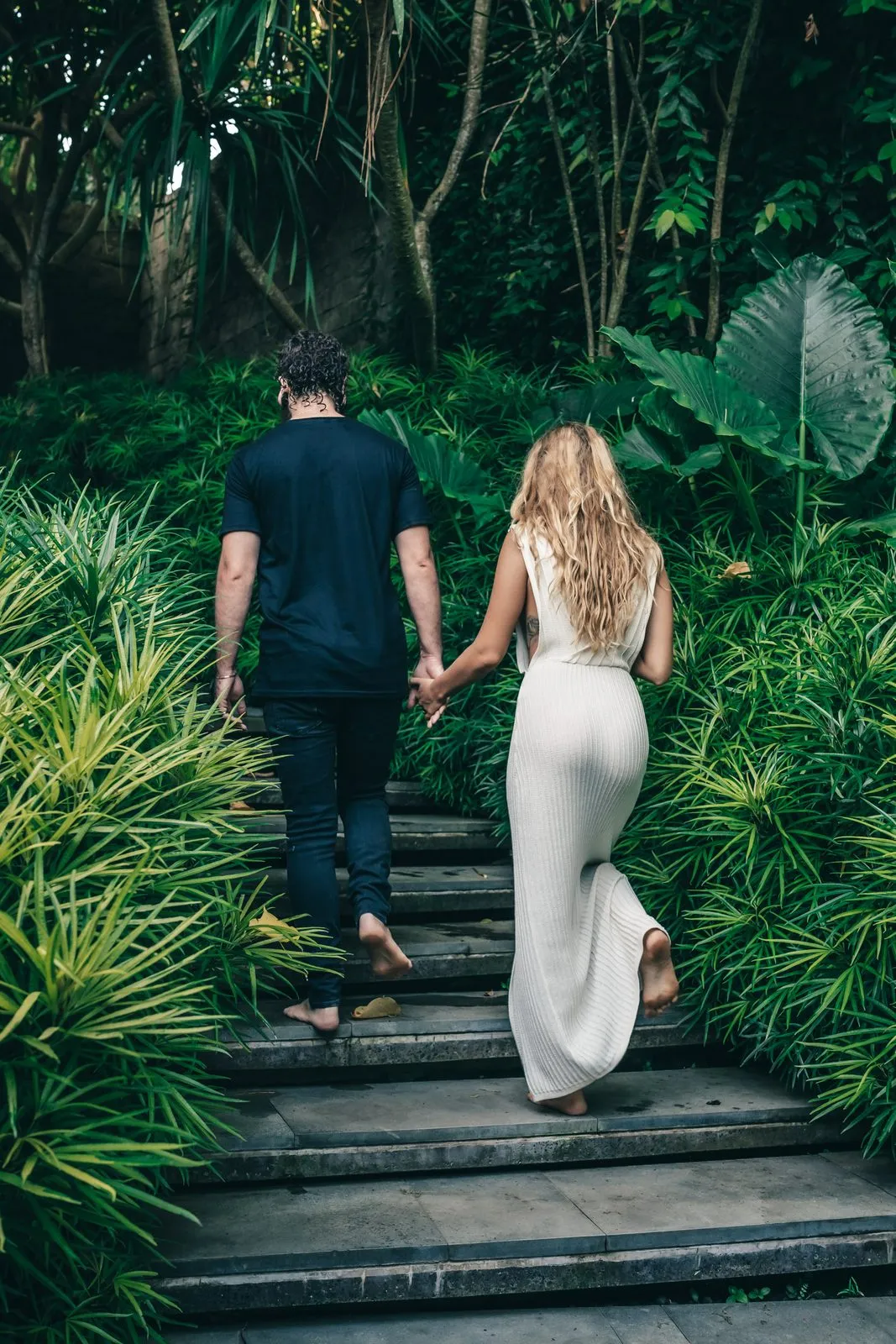 A couple climbing stone steps hand in hand through a jungle garden, seen from behind