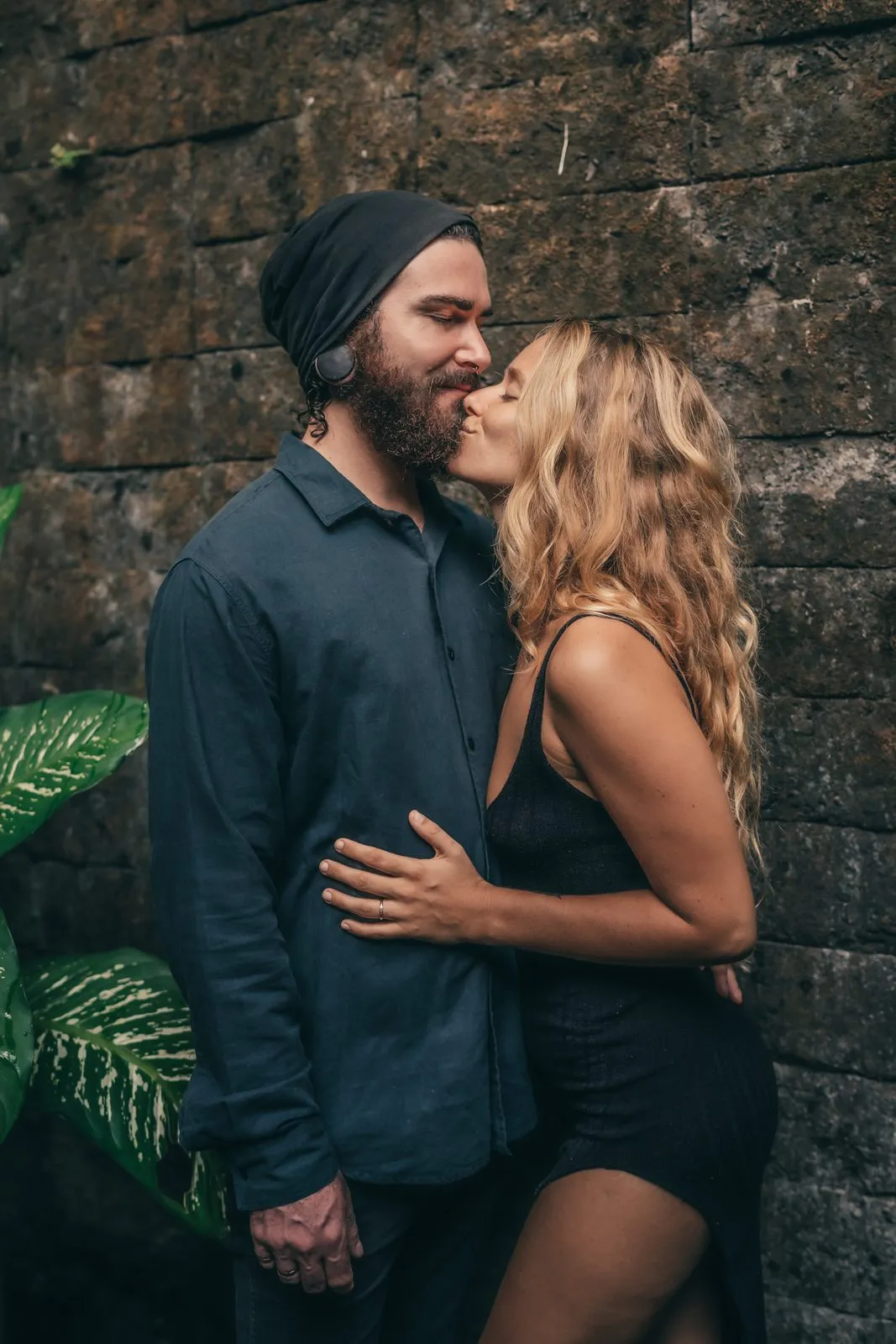 A laughing couple holding each other in front of a dark stone wall, he in a beanie