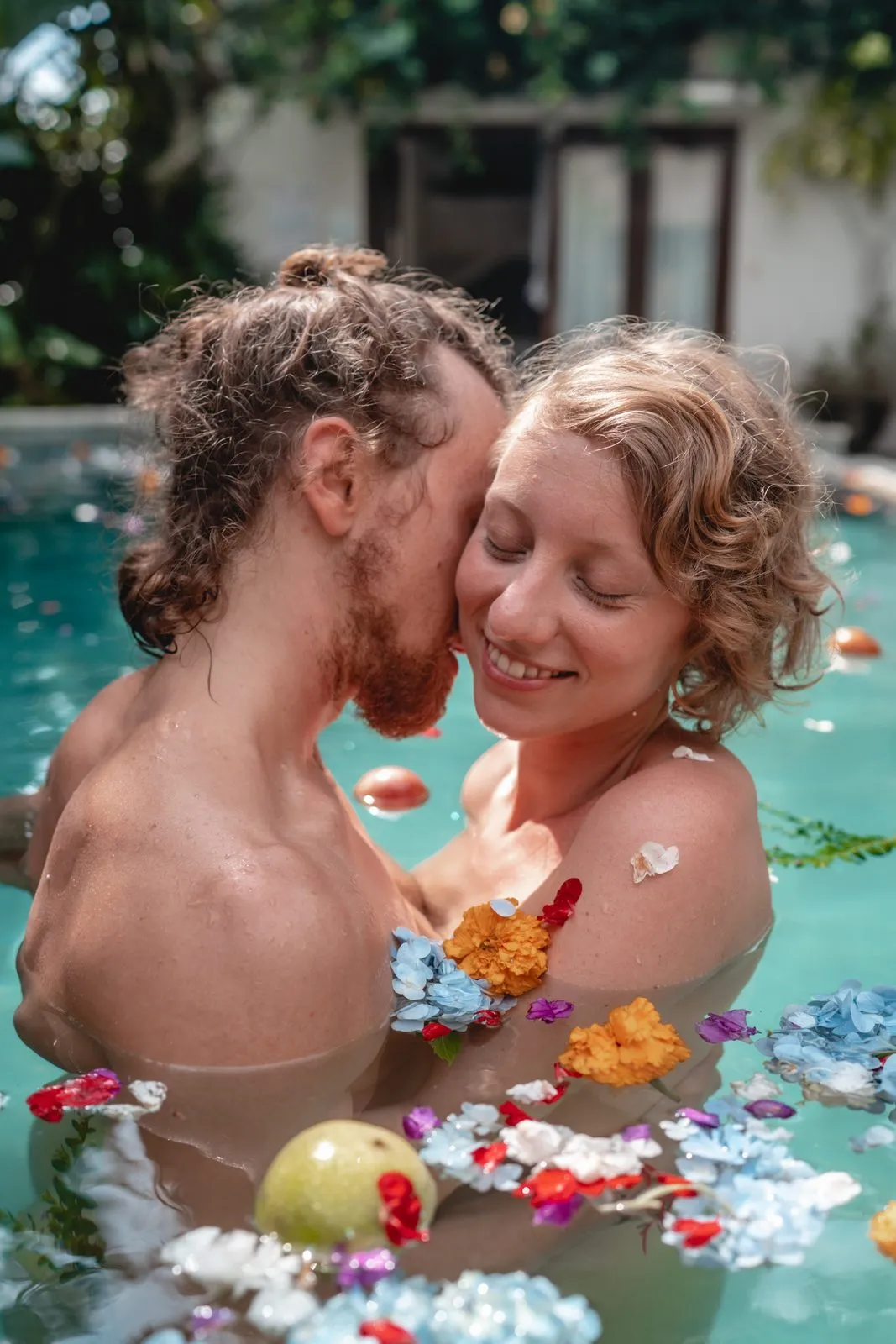 A couple in a villa pool among floating flowers and fruit, he kissing her cheek