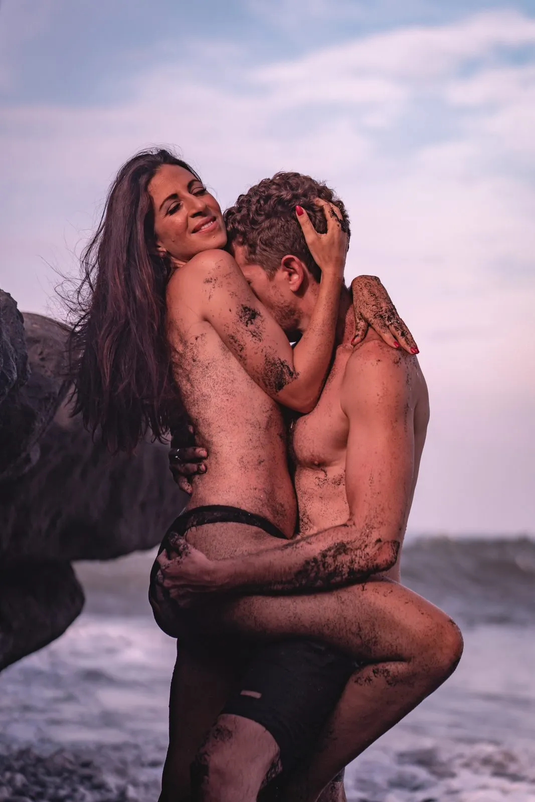 A couple embracing at the edge of the sea at dusk, wet sand on their skin, she laughing over his shoulder