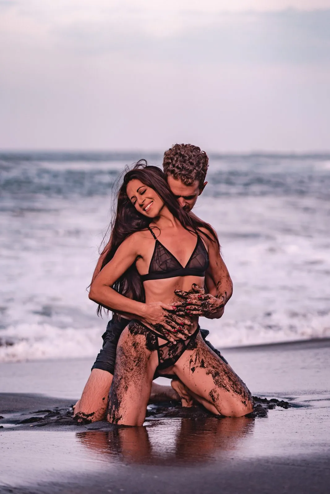 A couple kneeling in the surf on a black sand beach at sunset, his arms wrapped around her