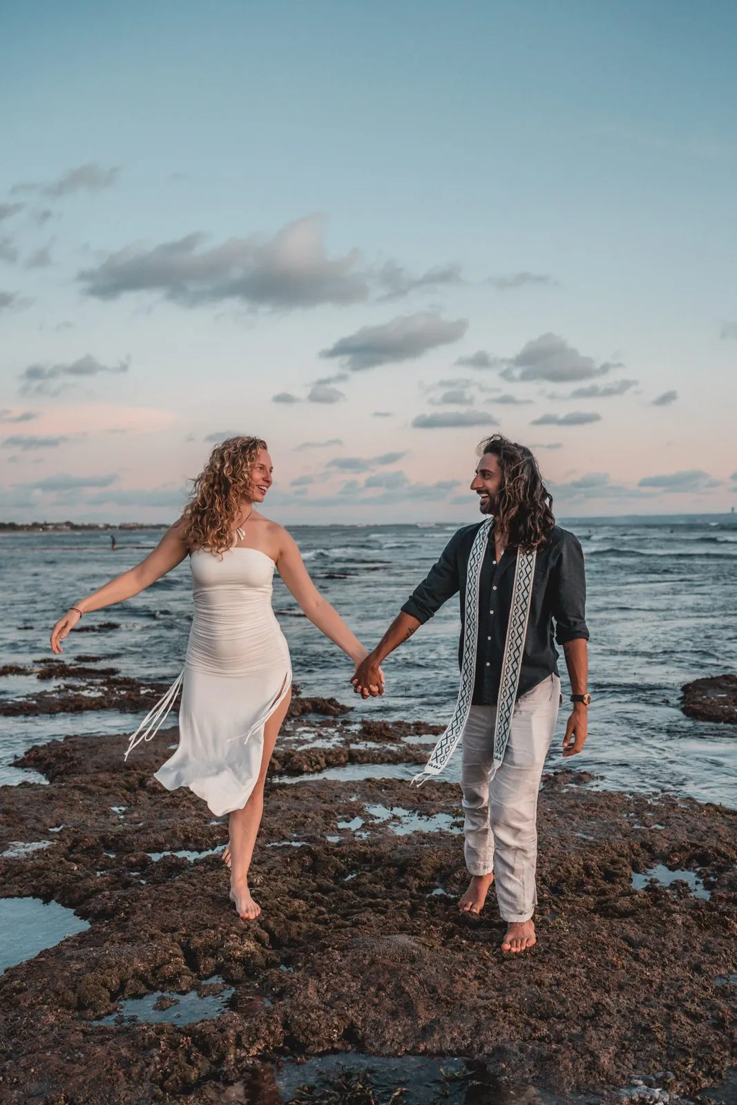 A couple holding hands across the rock pools at the shore at dusk, she in a white dress