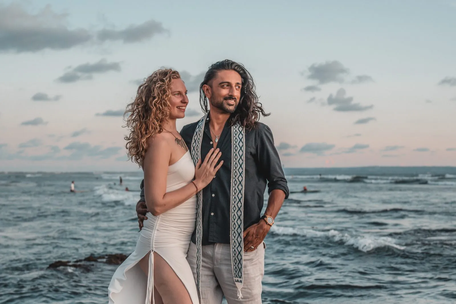 A couple standing together on a headland, her hand on his chest, the sea and a pale sky behind