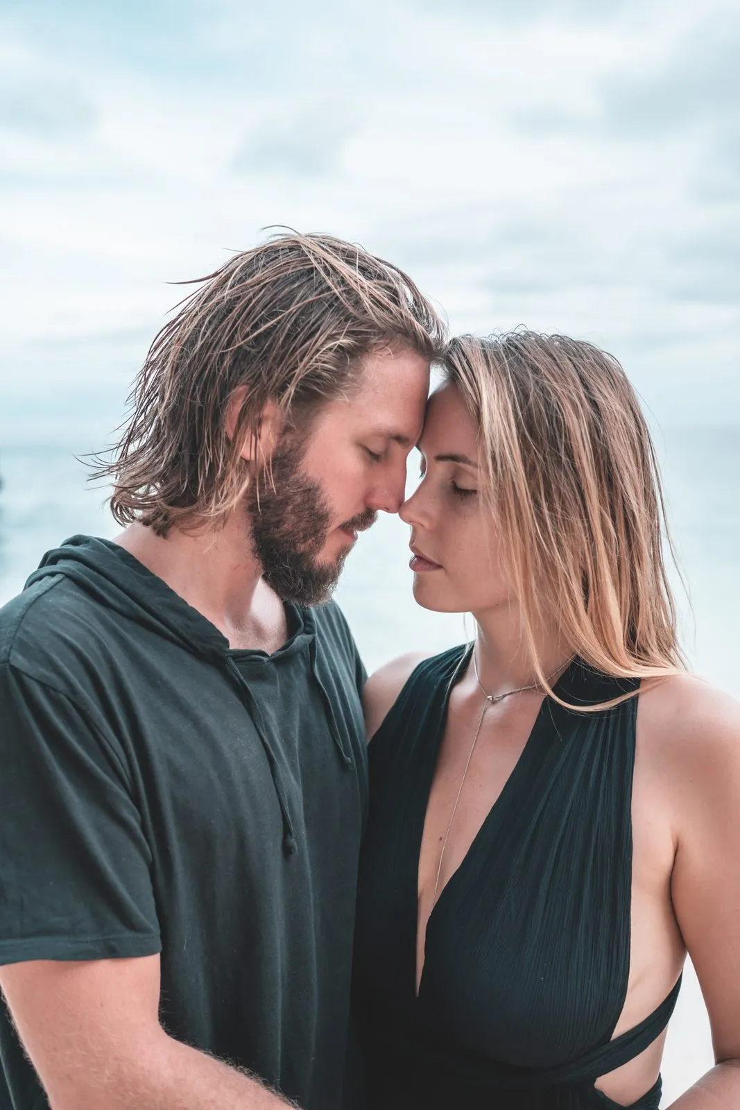 A couple nose to nose on the beach, she in a black dress, the sea behind them