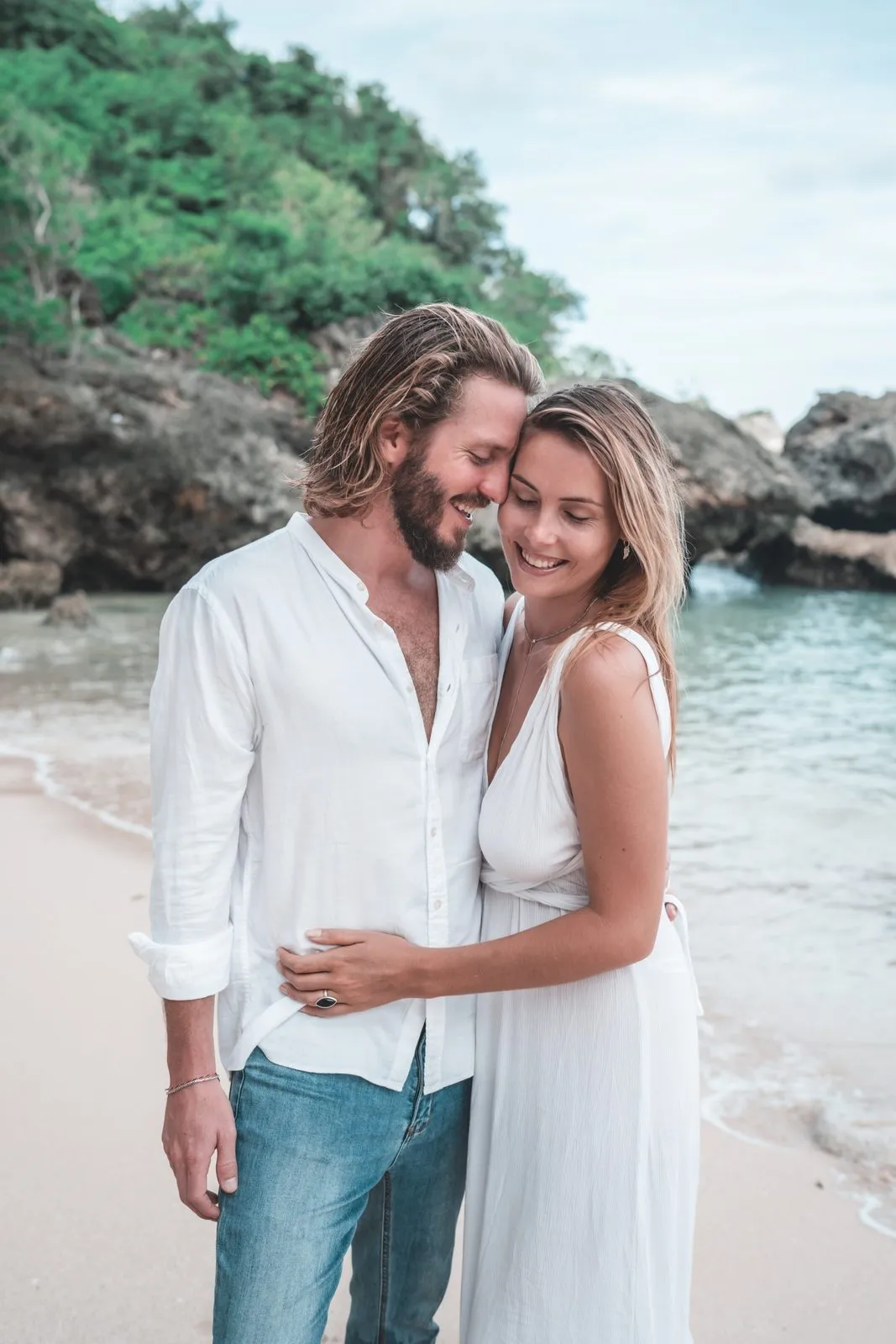 A couple in white embracing on a small beach at the foot of a cliff, the water turquoise behind
