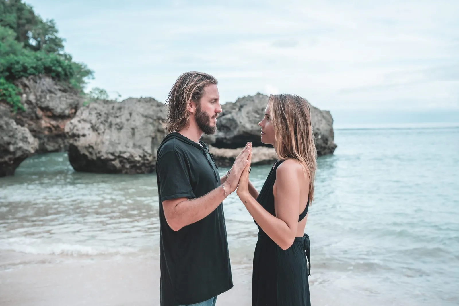A couple standing palm to palm in the shallows of a rocky cove