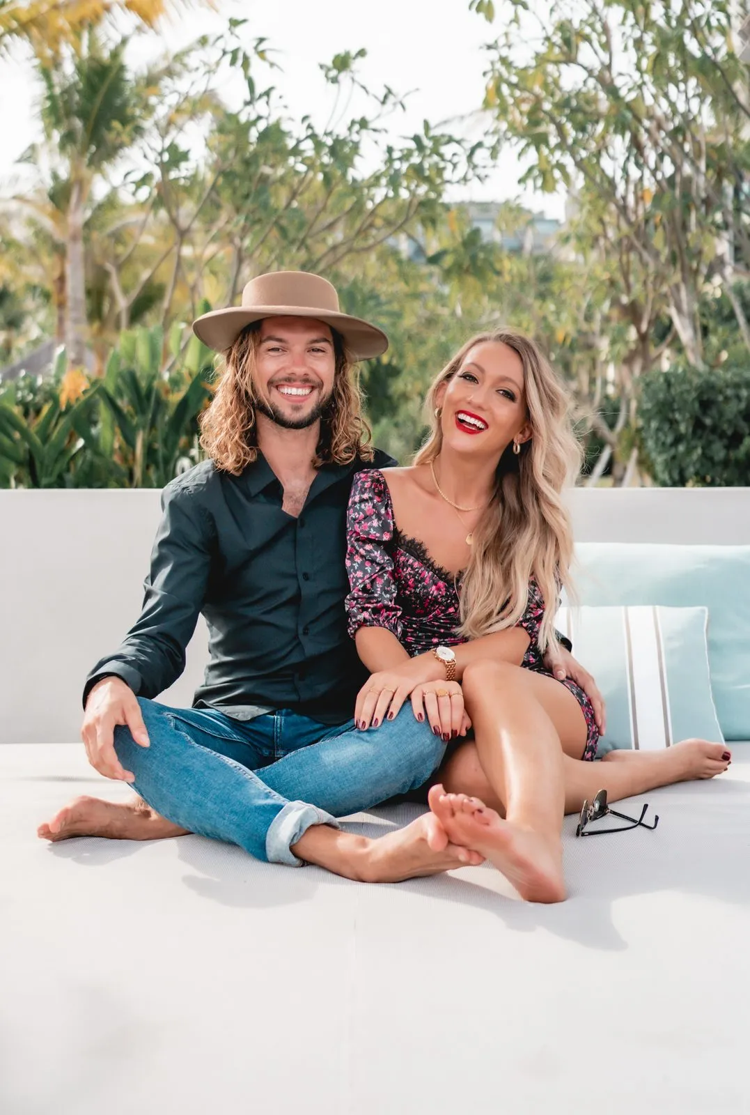 A couple sitting on a white daybed, he in a hat and denim, she leaning in, palms behind them