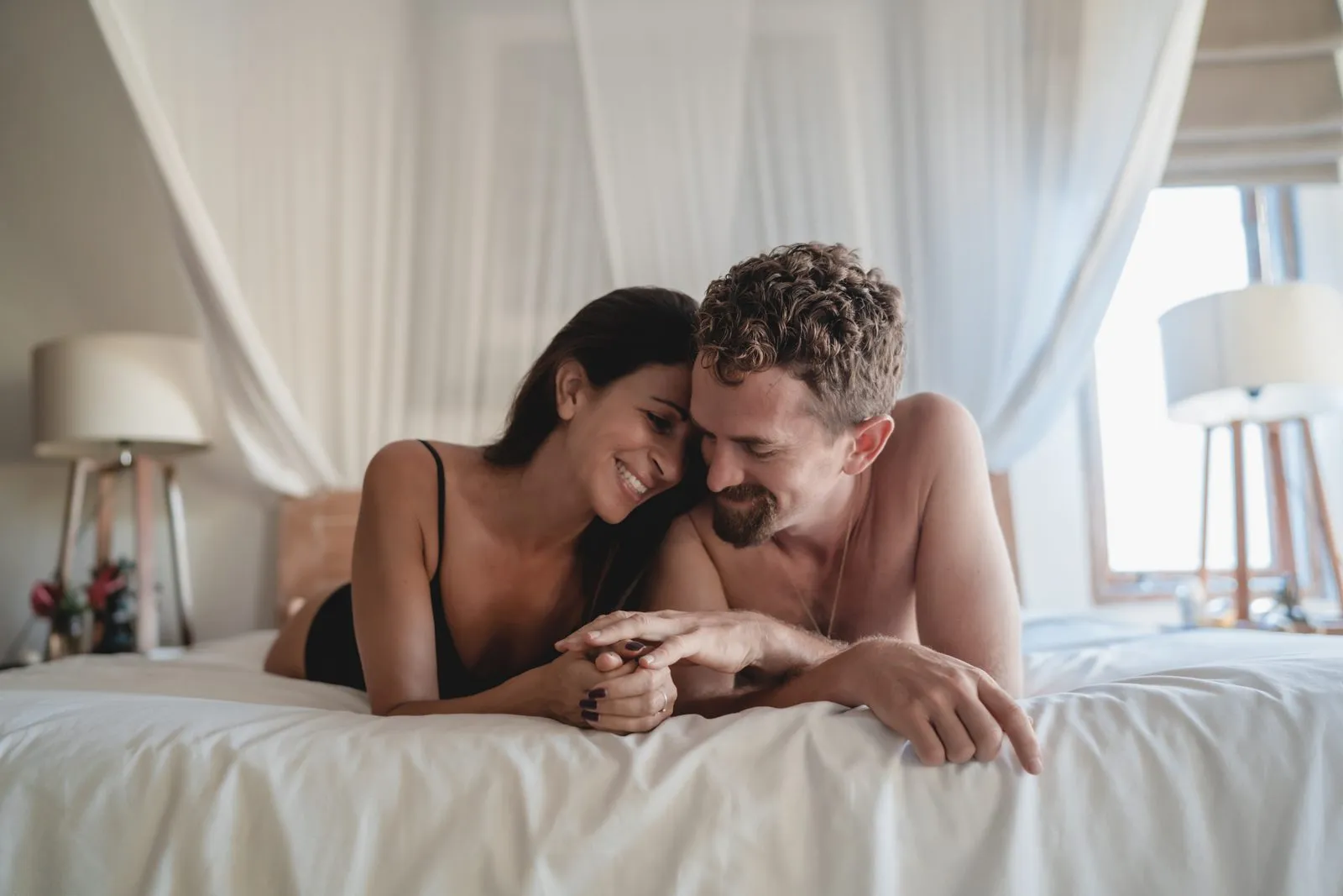 A couple lying on a white bed laughing, a canopy and soft light behind them