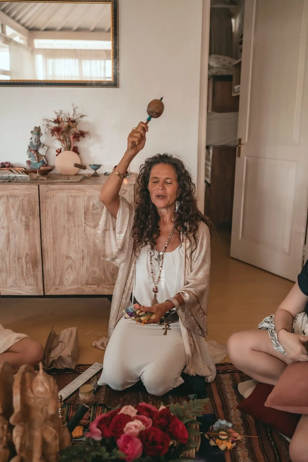 A woman in white kneeling with a rattle raised, leading a ceremony in a light-filled room