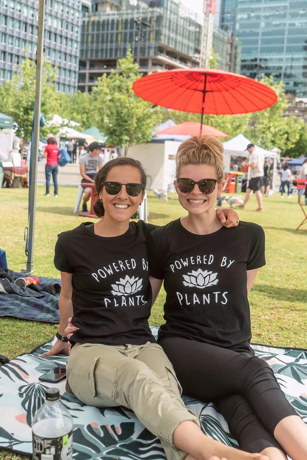 Two women in Powered by Plants T-shirts sitting on a picnic rug under a red parasol at the Adelaide Vegan Festival