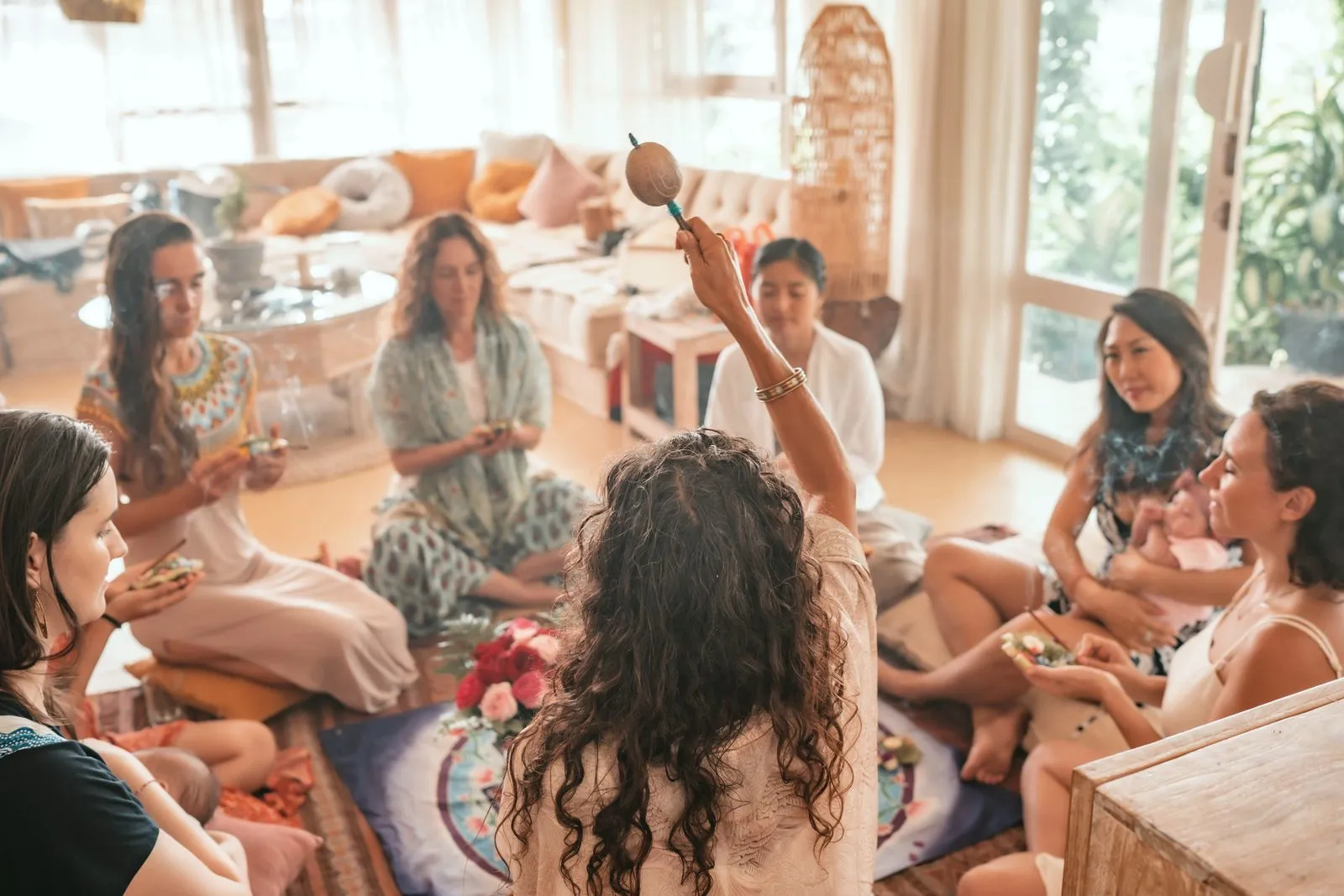 A circle of women on cushions in a bright room, one raising a rattle, at a women's gathering
