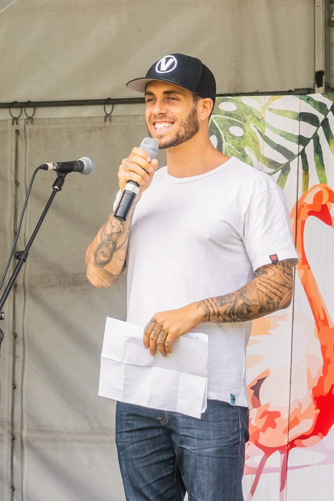 A speaker in a cap with a microphone on a festival stage, smiling, a flamingo banner behind him