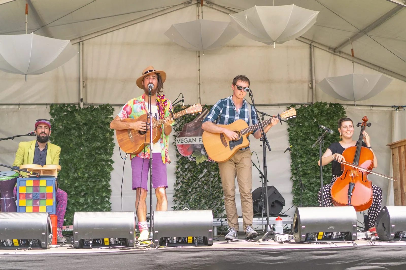 A band on an outdoor festival stage, singer, guitarist and cellist, under a white marquee