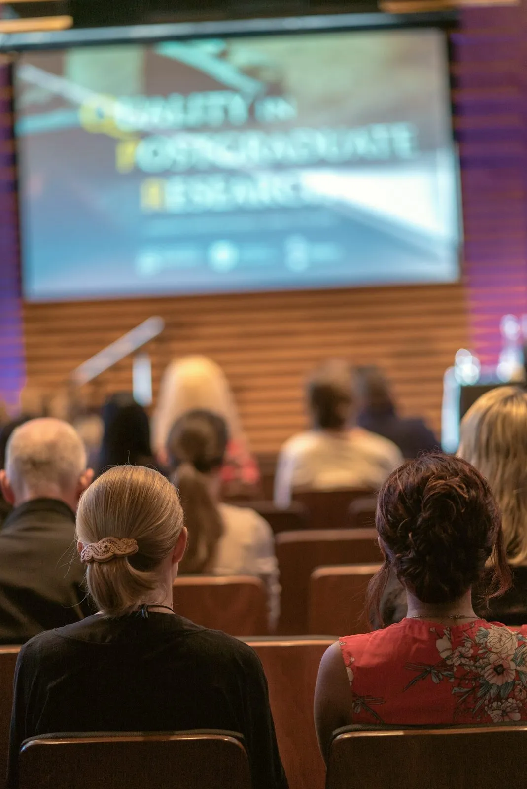 An audience seated at an academic conference, a presentation on the screen at the front