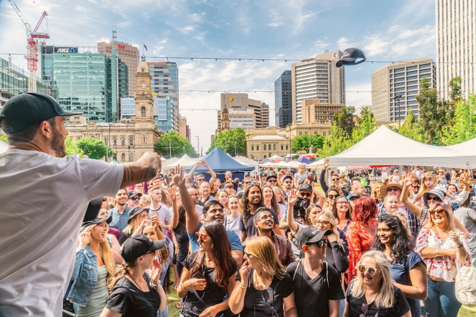 A crowd at an outdoor festival in a city square, an MC on stage in the foreground, Adelaide Vegan Festival