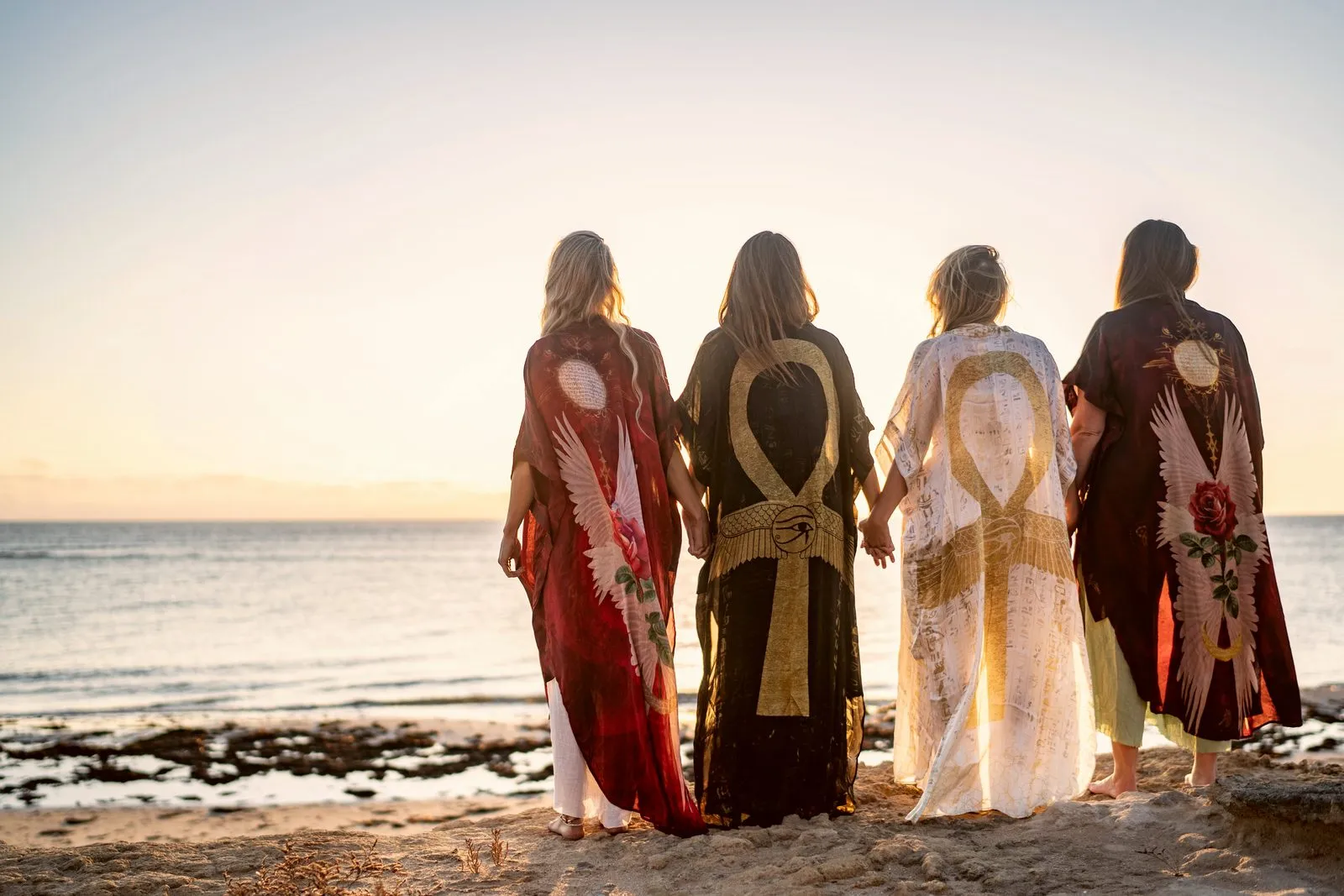 Four women in winged Nomadica kimonos standing with their backs to camera facing the sea at sunset, Australia