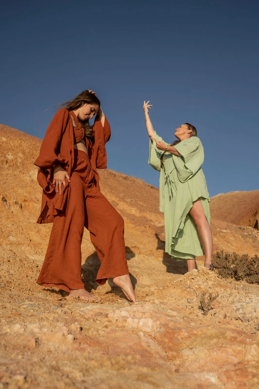 Two models in rust and sage linen against a deep blue sky on a red hillside, one reaching up, Nomadica