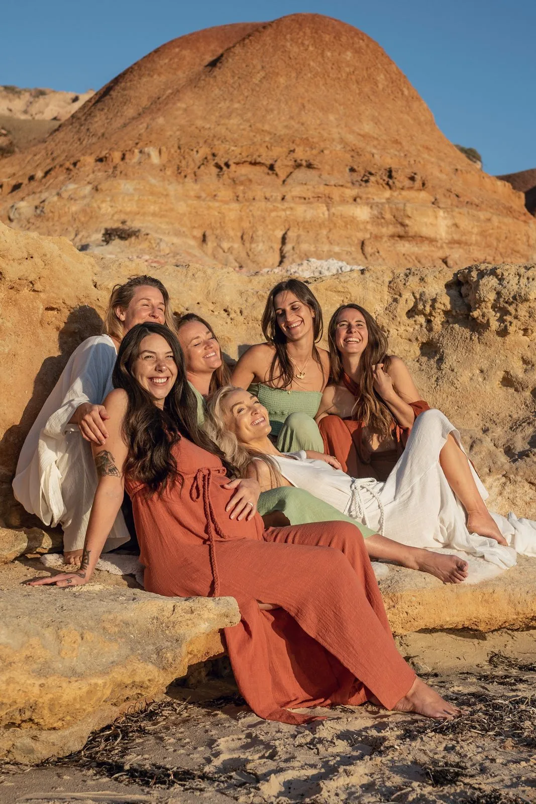 Six women in earth-toned linen sitting together on the rocks beneath red cliffs, Nomadica