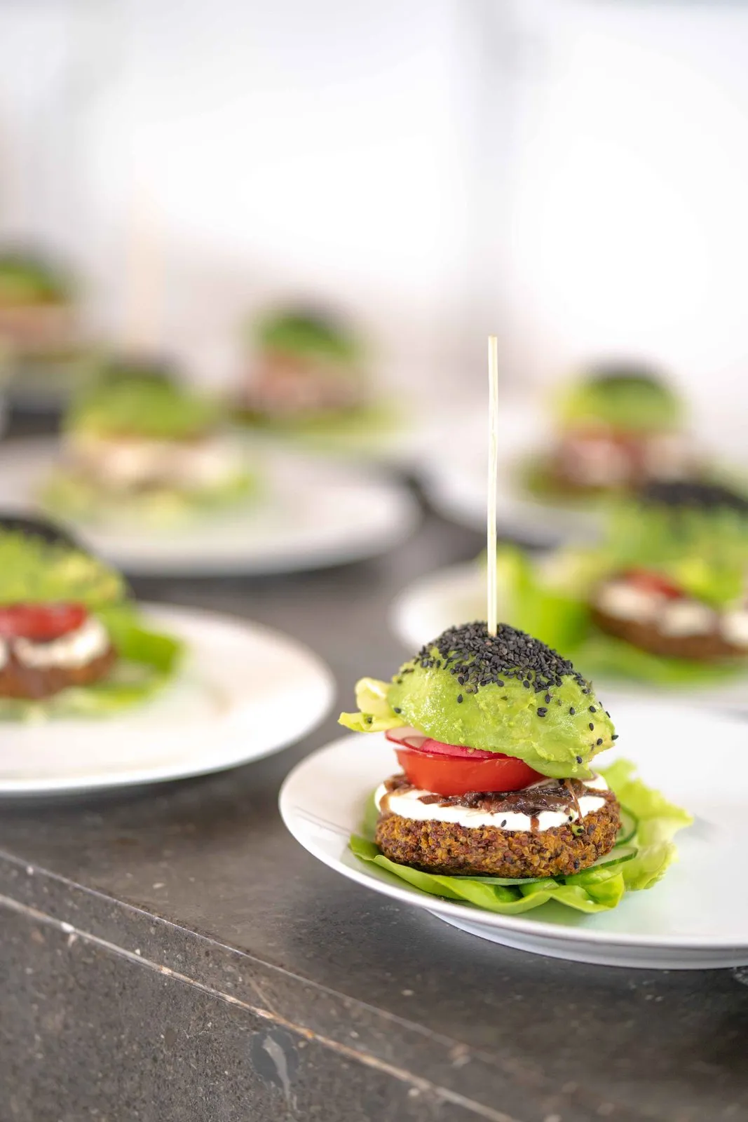 A row of plated vegan burgers with avocado and tomato, each held with a skewer, on a restaurant pass