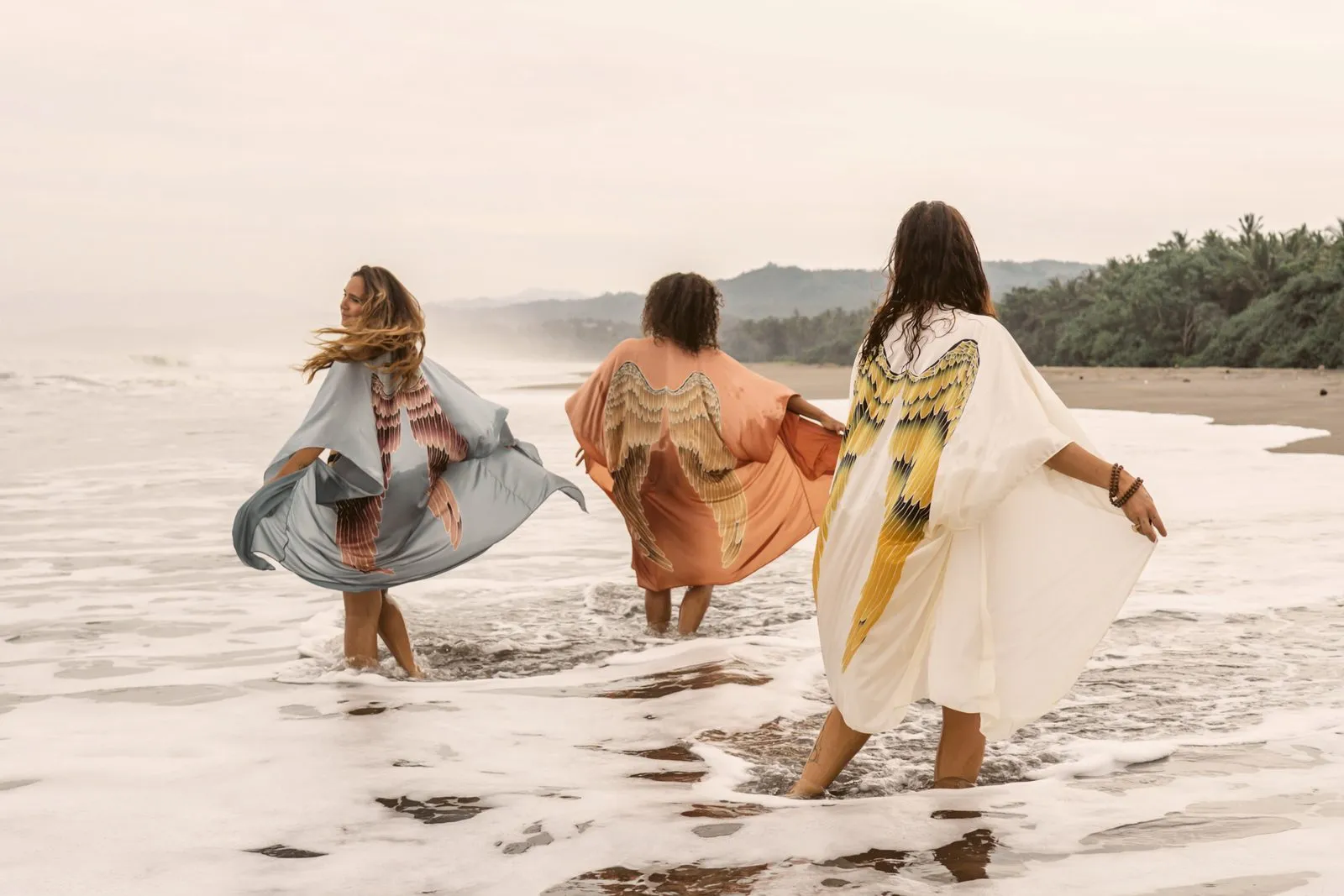 Three women in feathered kimonos running through the surf, jungle behind the beach, Warriors of the Divine