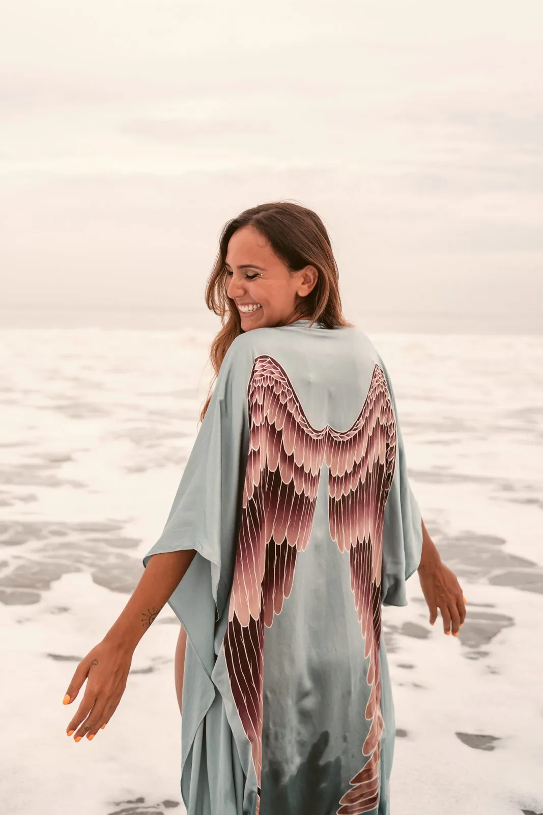 A smiling woman in a pale blue kimono printed with wings, seen from behind on the beach, Warriors of the Divine