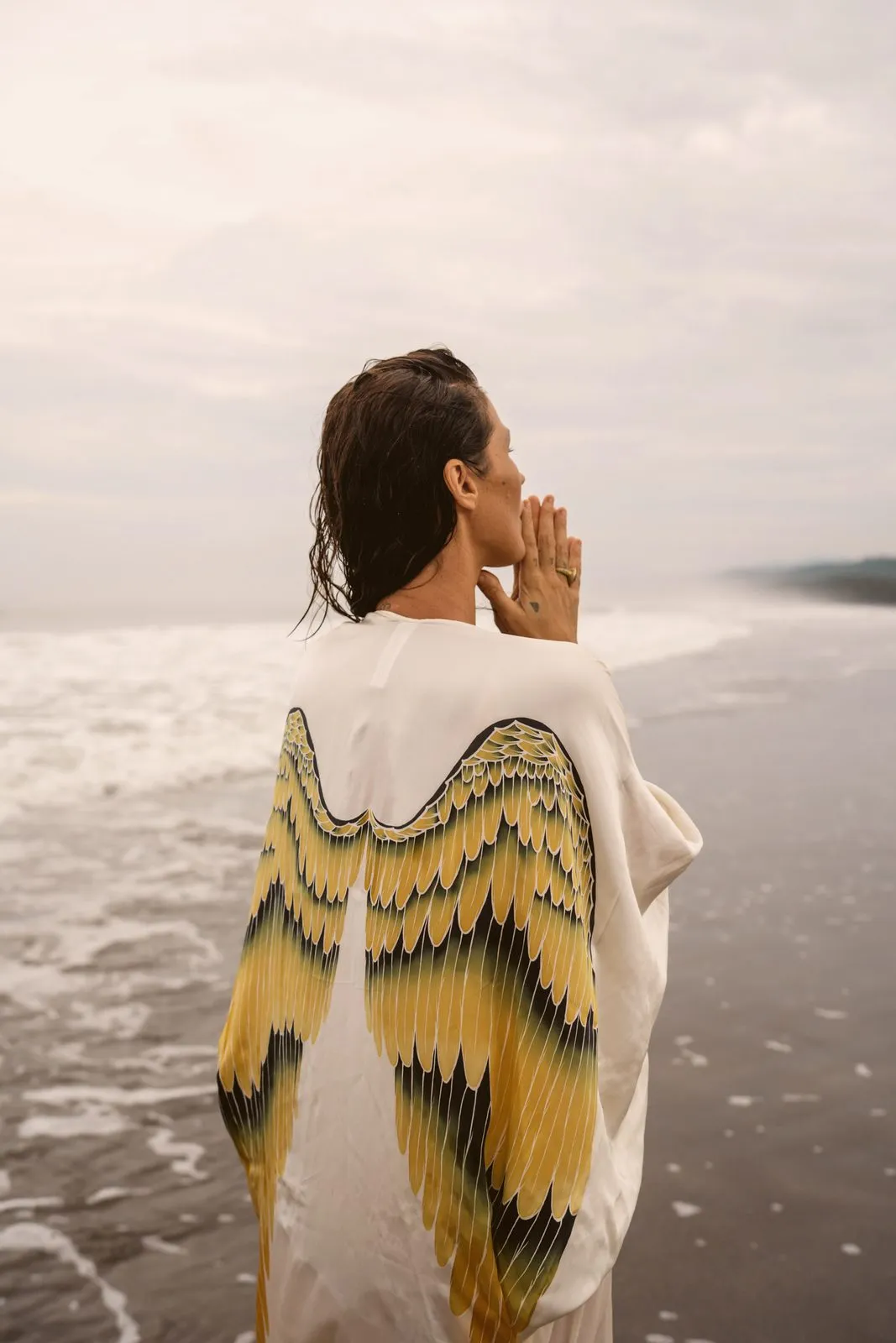A woman in a white kimono with yellow feathered wings, seen from behind facing the sea, Warriors of the Divine