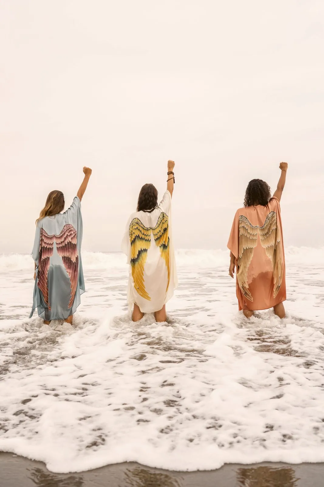 Three women in winged kimonos standing in the surf with their fists raised, backs to camera, Warriors of the Divine