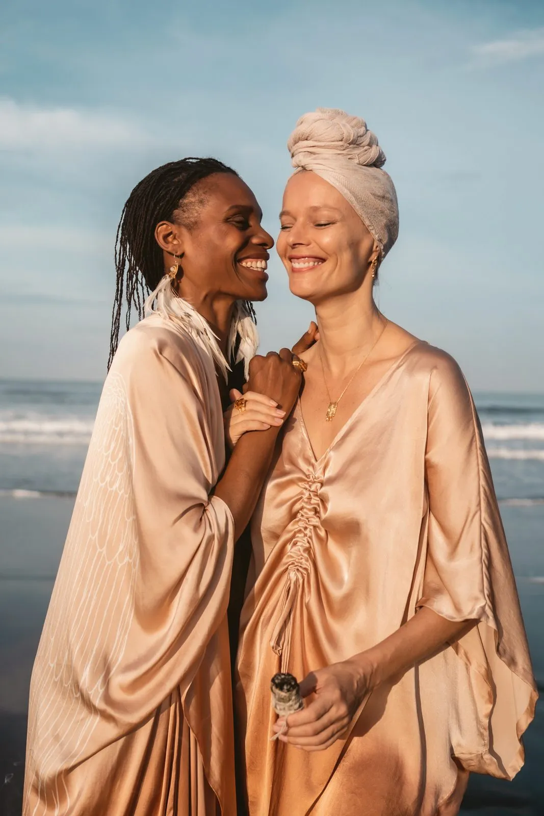 Two women in champagne satin kimonos laughing together on the beach, one in a head wrap, Warriors of the Divine