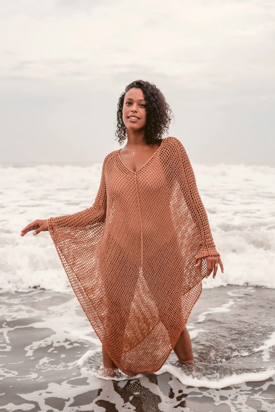 A woman in a rust crochet kaftan standing in the surf, Warriors of the Divine