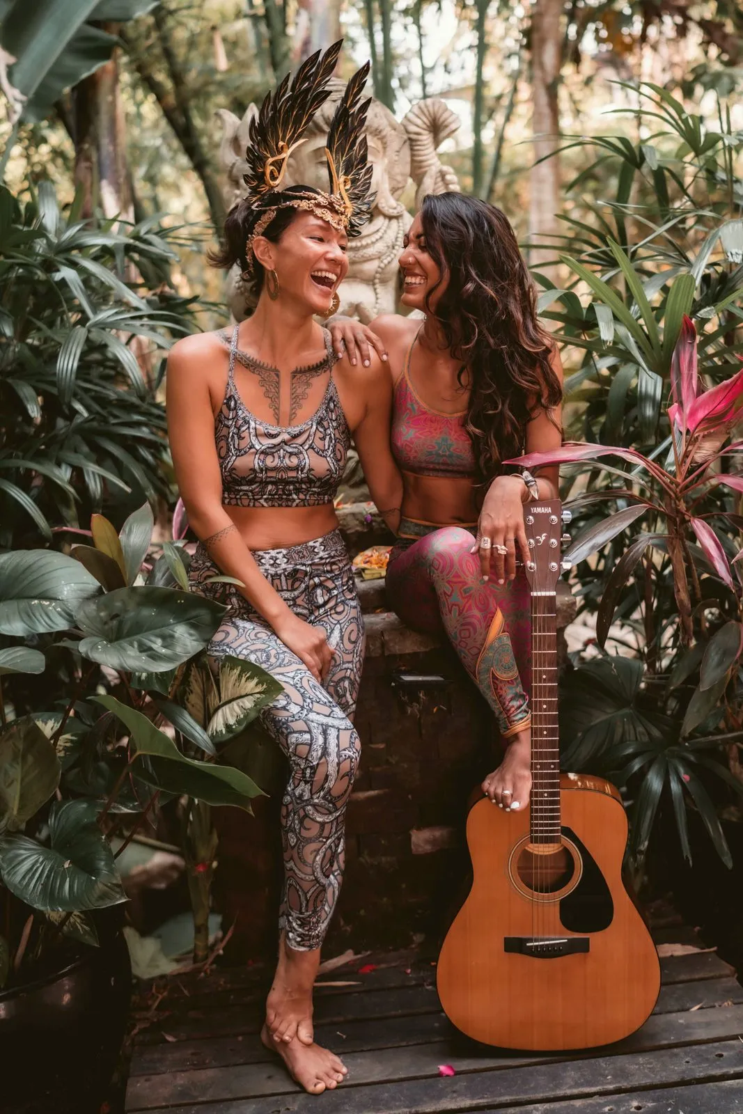 Two women in feathered headdresses and Miramaya activewear laughing together with a guitar, in a garden