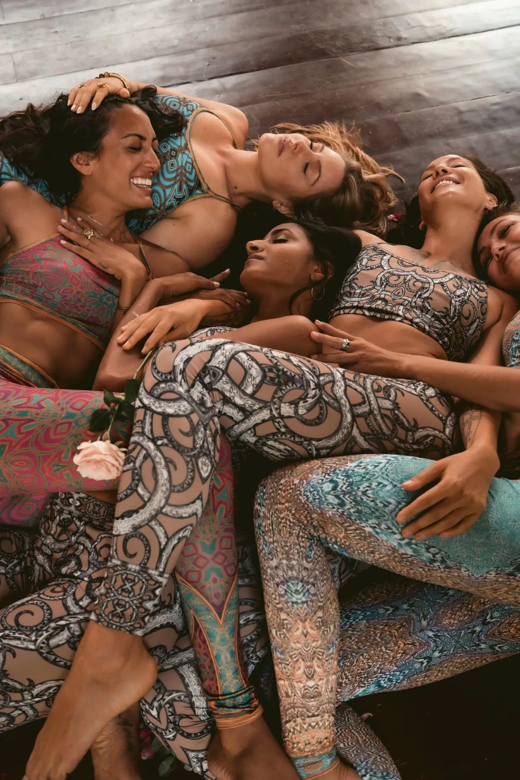 Four women in patterned Miramaya activewear lying together on a wooden deck, laughing, photographed from above